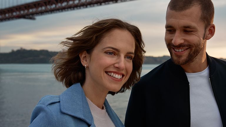 Smiling woman and smiling man standing next to each other, in the background is a bridge over a body of water